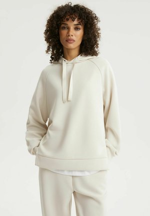 Kobieta o kręconych włosach, ubrana w oversize'ową kremową bluzę z kapturem i dopasowane kremowe spodnie, stojąca z rękami w kieszeniach na jednolitym tle.