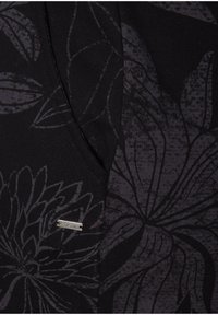 Svart plagg med ett blommönster i mörkgrått. Har en V-ringning och en metallisk varumärkesetikett. Mjuk textur med detaljerade tryck.