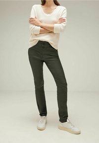 Ljusbeige stickad tröja med V-hals, tillsammans med mörkgröna slim-fit byxor med knappstängning. Modellen bär vita sneakers.