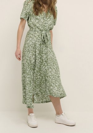 Woman wearing a green floral midi dress with buttons and tie waist, paired with white sneakers, standing against a plain background.