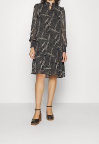 Vestido negro con patrón de olas, mangas largas y cuello con abertura. Presenta puños fruncidos y un dobladillo en capas. Se lleva con zapatos de tacón bloque negro.