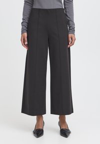 Kobieta ubrana w czarne, szerokie spodnie o długości do połowy łydki, szary top z długim rękawem oraz czarne buty na spiczastym nosku z ozdobą w postaci podwójnej klamry.