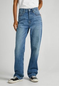 High-waisted, loose-fit blue jeans made from denim, featuring minimal fading, five pockets, and a button closure. Worn with black sneakers.