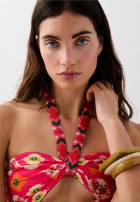 Giovane donna con lunghi capelli scuri indossa un top halter rosso a fantasia, una collana di perline e due bracciali spessi al polso, su uno sfondo neutro.