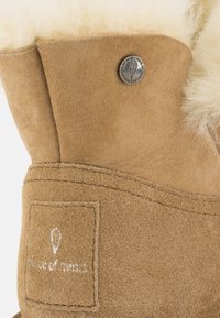 Stivale in suede beige con un morbido bordo in pelliccia di shearling crema, etichetta con logo in rilievo e un dettaglio in metallo argentato con bottone sul lato superiore.