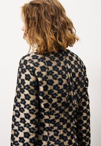 Dierenprint jas met een gestructureerde beige stof en zwarte bloemenpatronen, een getailleerd ontwerp en een achterkant met een plooi-detail.