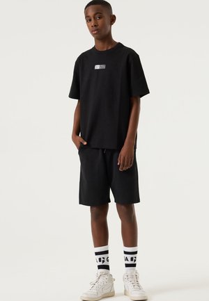 T-shirt en coton noir à manches courtes avec un logo imprimé. Assorti à un short noir, des baskets montantes blanches et des chaussettes rayées.