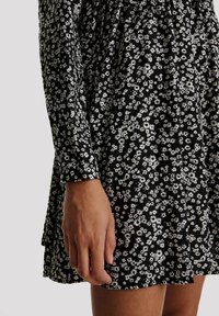 Schwarzes Kleid mit einem weißen Blumenmuster, langen Ärmeln, gerafftem Stoff im Oberteil und einem ausgestellten Rockdesign.