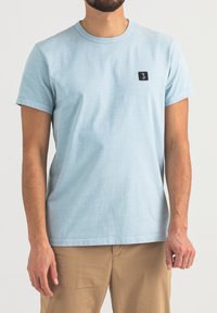 Helles blaues T-Shirt aus weichem Stoff mit Rundhalsausschnitt und kurzen Ärmeln. Verfügt über ein kleines schwarzes quadratisches Logo auf der linken Brustseite.