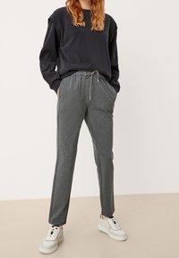 Mujer vestida con pantalones de chándal oscuros, parte superior negra de manga larga y zapatillas blancas, de pie con las manos en los bolsillos sobre un fondo neutro.