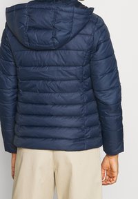 Tmavě modrá prošívaná péřová bunda s kapucí, s prošíváním, horizontálním šitím a elastickými manžetami. Materiál vypadá hladce a je izolovaný.