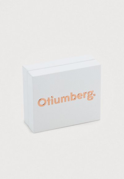 Λευκό ορθογώνιο κουτί με υφή, με τη λέξη "Otiumberg" σε ανάγλυφα γράμματα από ροζ χρυσό στο μπροστινό μέρος.