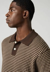 Ruskea ja beige houndstooth-mallinen polo-paita, jossa on ruskea kaulus, kolme napin aukkoa ja teksturoitu neulottu kangas.