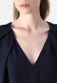 Abito blu navy con scollo a V profondo e dettaglio delle spalle drappeggiato. Texture liscia, cuciture minime ed elegante silhouette.