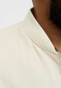 Light beige bomber jacket with a textured fabric, featuring a ribbed collar and a zipper closure, close-up of the collar area.