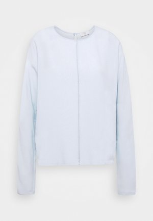 Chemise bleu clair à manches longues avec un col rond et une couture centrale visible courant verticalement sur le devant.