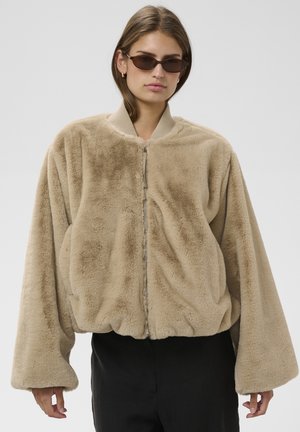 Vrouw draagt een oversized beige kunstbontjas met rits en zwarte broek, gecombineerd met kleine donkere zonnebrillen en oorbellen in hoepelvorm.