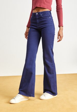 Jean bleu marine évasés en denim avec une texture lisse, dotés d'une fermeture à bouton et de poches avant, accompagnés de tennis blanches.