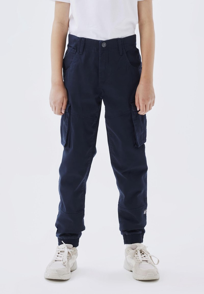 Enfant portant un pantalon cargo bleu marine avec des poignets élastiques et des baskets blanches, debout devant un fond uni clair.