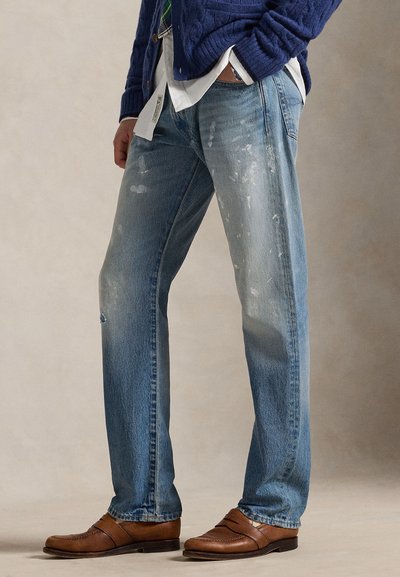 Homme portant un jean bleu clair éclaboussé de peinture, mocassins en cuir marron, chemise blanche, pull bleu marine, mains dans les poches, debout sur un fond neutre.