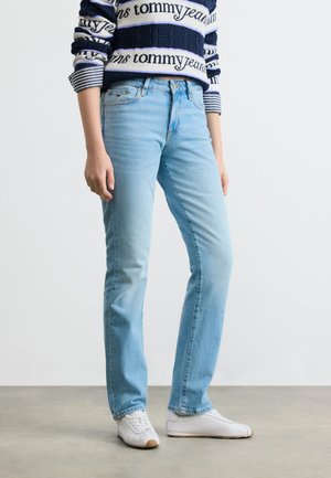 Osoba ve světle modrých džínech rovného střihu, bílých teniskách a modrobíle pruhovaném svetru Tommy Jeans stojící na šedé podlaze.