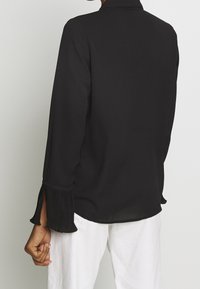 Blouse noire à manches longues avec des accents plissés aux poignets, un col et un tissu léger et lisse.