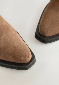 Botas de ante marrón hasta el tobillo con punta afilada y suela de goma negra. La textura es suave, con un diseño limpio y minimalista.