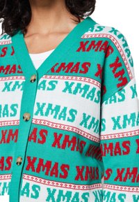 Sweter z dzianiny z turkusowym kołnierzem i guzikami, z wyraźnymi czerwonymi i zielonymi wzorami tekstu "XMAS" na białym tle.