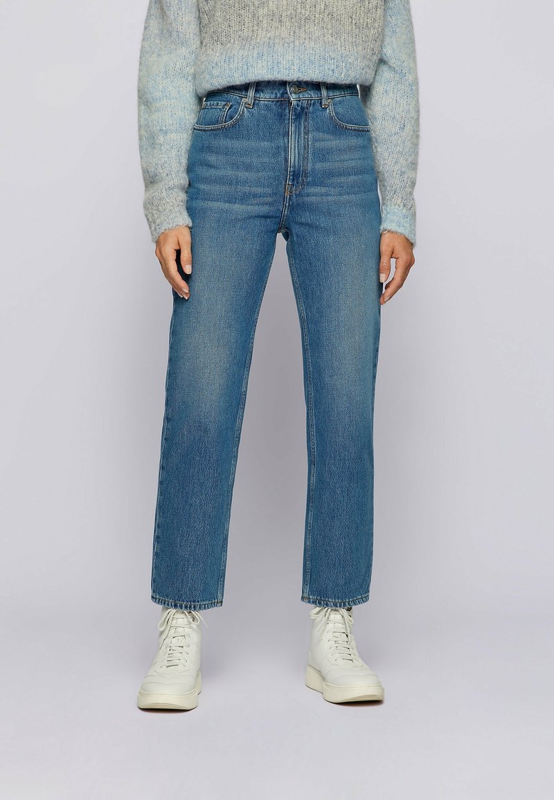 Jean taille haute à jambes droites en denim bleu moyen. Caractérisé par un ourlet effiloché et cinq poches. Associé à des baskets blanches et un pull gris.