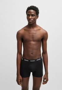 Schwarze Boxer Shorts mit einem elastischen Bund, der "HUGO" zeigt. Das Material wirkt glatt und eng anliegend, mit einem mittelhohen Schnitt.