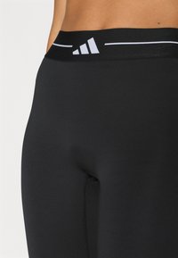 Leggings de sport noirs avec une taille haute ornée de bandes blanches et un logo. Le tissu est lisse avec une légère brillance.