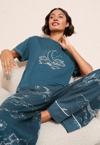 Tirkīza viesnīcas komplekts, kas sastāv no īsām piedurknēm t-krekla ar mēness un mākoņu dizainu, un saskaņotiem biksēm ar debesu rakstiem un baltu apdari.