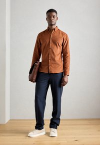 Cămașă maro cu nasturi dintr-un material moale, asociată cu pantaloni negri închis și adidași albi. Ținând o geantă maro tip messenger, stând pe un podea din lemn.