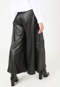 Schwarze, fauxlederne, weit geschnittene Hosen mit strukturierter Oberfläche, ausgestattet mit einer seitlichen Tasche, die mit einem Knopf gesichert ist. Kombiniert mit kniehohen, strukturierten Stiefeln.