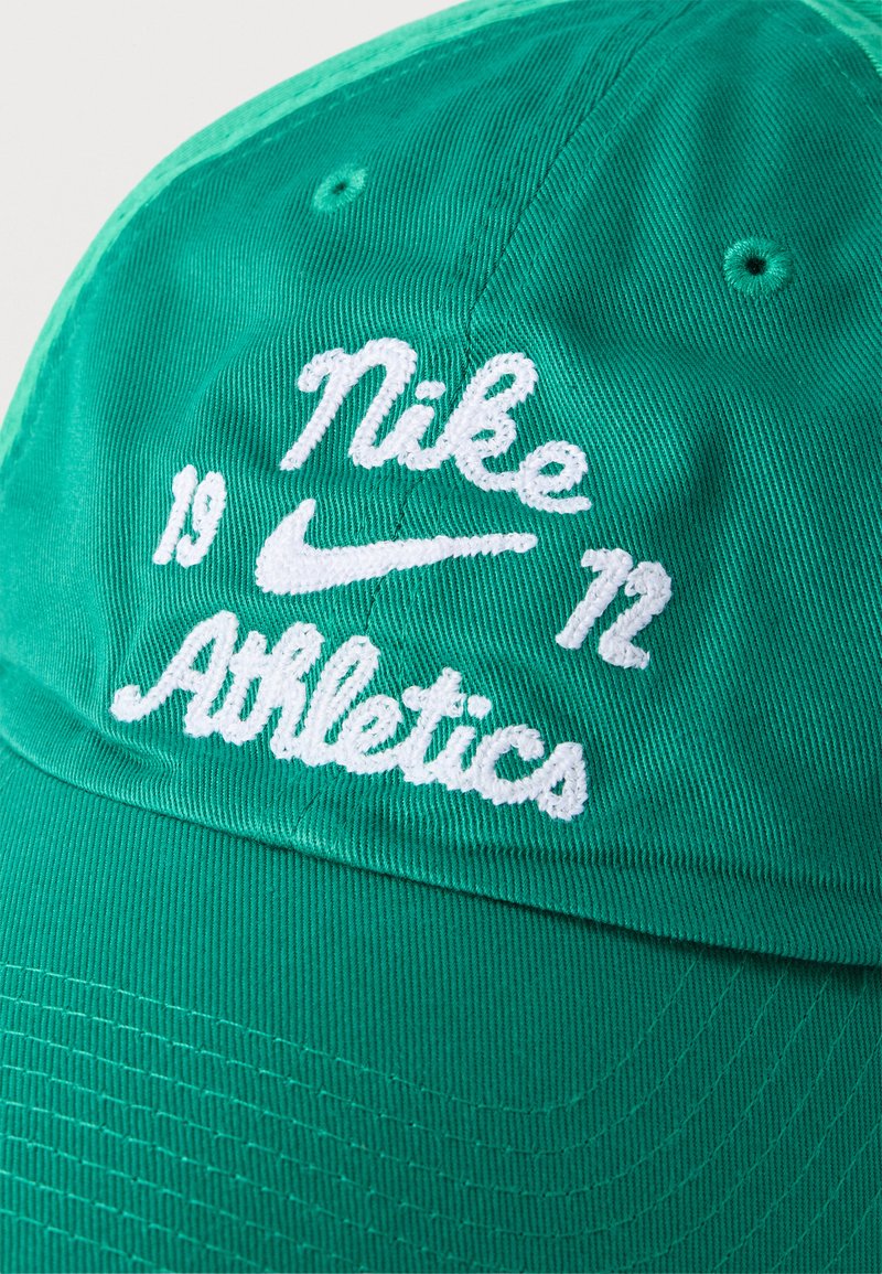 Grøn stofkasket med hvidt broderet tekst "Nike Athletics" og numrene "19" og "72" på hver side af det hvide Nike swoosh-logo.