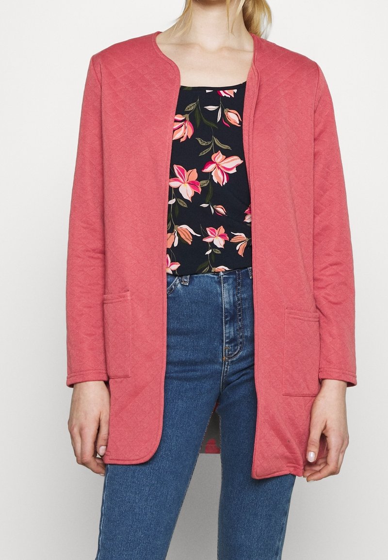 Femme portant une veste matelassée corail ouverte sur le devant, au-dessus d’un top noir fleuri et d’un jean bleu, debout les bras détendus le long du corps.
