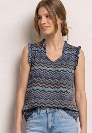 Femme portant une blouse sans manches à motif zigzag dans des tons bleus, marron et blancs, associée à un jean bleu clair, debout contre un mur texturé.