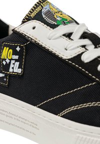 Baskets noires en toile avec des lacets blancs et une semelle en caoutchouc. Étiquette jaune et noire avec le texte "Mother" et un design graphique sur le côté.