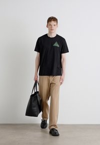 Svart t-skjorte med korte ermer og grønt geometrisk design på venstre bryst, kombinert med beige bukser og bærene en svart totebag.