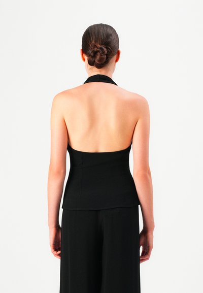 Woman with dark hair in a braided bun wearing a black halter top and black pants, viewed from the back against a white background.