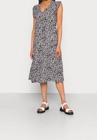 Blumenkleid in Midi-Länge mit kurzen Rüschenärmeln, V-Ausschnitt, navy Hintergrund und Muster aus weißen/rosa Blumen. Kombiniert mit weißen Sandalen.