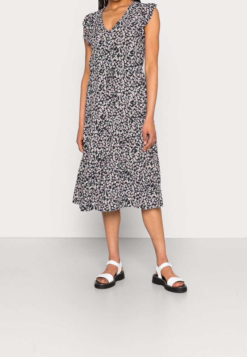 Blumenkleid in Midi-Länge mit kurzen Rüschenärmeln, V-Ausschnitt, navy Hintergrund und Muster aus weißen/rosa Blumen. Kombiniert mit weißen Sandalen.