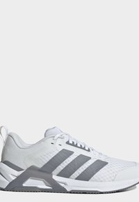 Chaussure de sport blanche avec des bandes grises, dessus en mesh, semelle rembourrée et lacets sur un fond clair uni.
