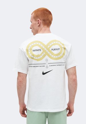 Άτομο με κόκκινα μαλλιά, ορατό από πίσω, που φοράει λευκό μπλουζάκι με κίτρινο γραφικό άπειρου δρόμου και κείμενο "Infinite Pursuit" και λογότυπο της Nike.