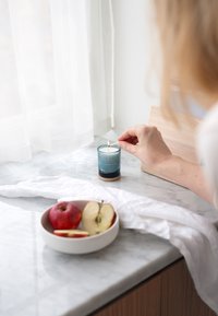 Blaue Glaskerze mit Holzunterlage, die auf einer marmorierten Arbeitsplatte brennt, daneben eine Schüssel mit roten und grünen Apfelscheiben. Im Hintergrund ein weißer Vorhang.