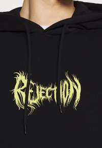 Primo piano di una felpa nera con cappuccio con testo giallo in stile fiamma che recita "Reject Only" nella zona del petto.