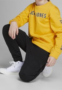 Żółta bluza z nadrukiem "JCK JONES", w połączeniu z czarnymi spodniami cargo z bocznymi kieszeniami i elastycznymi mankietami, noszona z białymi trampkami.