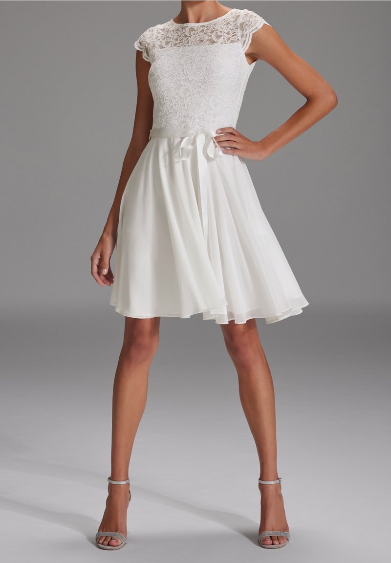 Femme portant une robe blanche sans manches avec un corsage en dentelle, une taille ajustée nouée avec un ruban, et une jupe fluide longueur genou, associée à des talons ouverts.