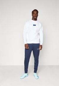 Sweatshirt blanc avec une étiquette rectangulaire marine portant le mot "PARIS", accompagné de joggings marins et de chaussures de sport bleu clair.