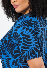 Camiseta azul con un patrón de hojas negras, de manga corta, cuello redondo y textura de algodón suave. La vista lateral muestra claramente el diseño.